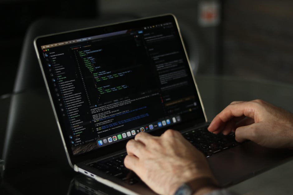 A close-up view of a person's hands typing on a laptop with a dark screen displaying lines of code in various colours, situated on a black table or desk. The person's fingers are positioned over the k